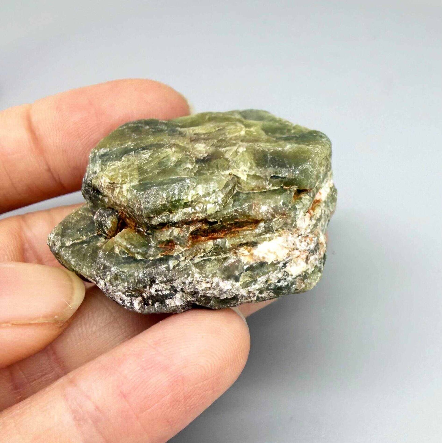 Natural rough green sapphire, opaque, uncut and irregular, showing olive to moss-green tones, photographed in hand on neutral background.