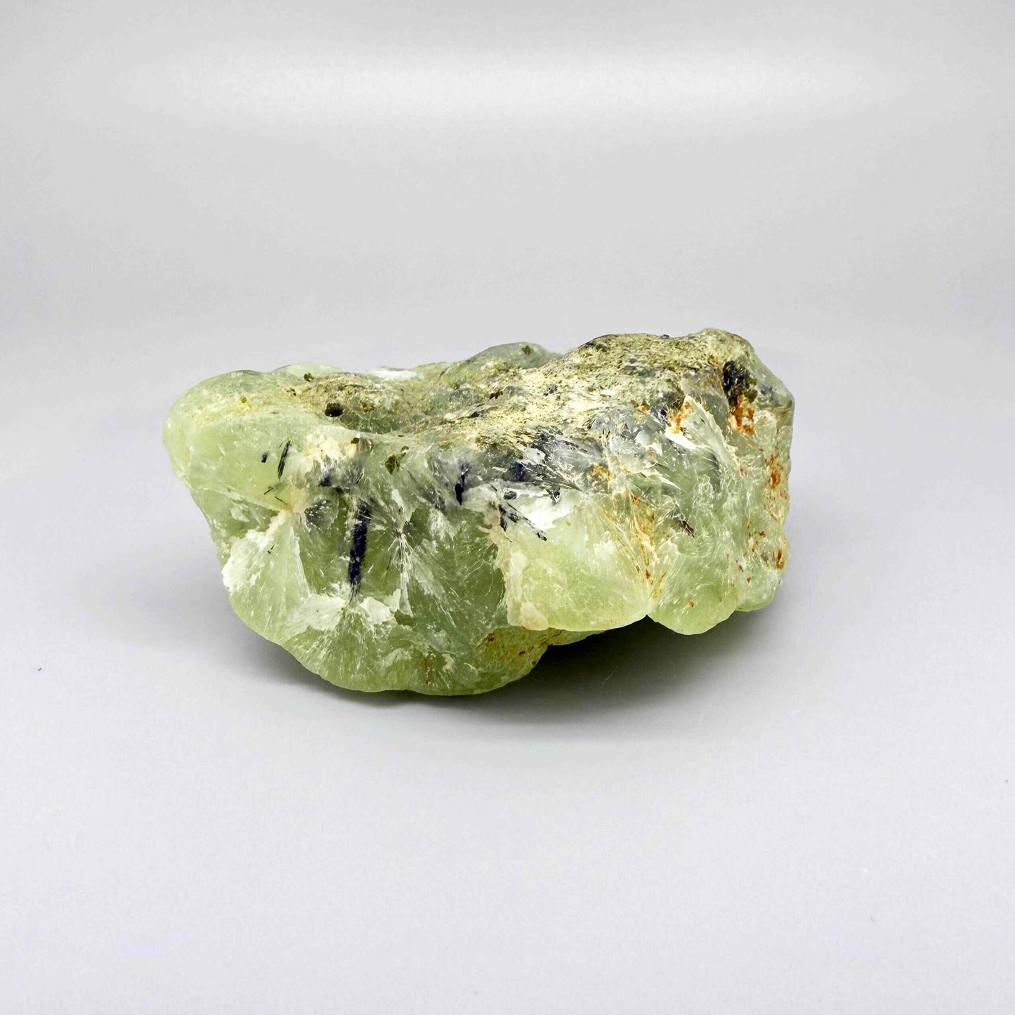 Prehnite rough stone in pale apple-green color with dark inclusions and semi-translucent body.