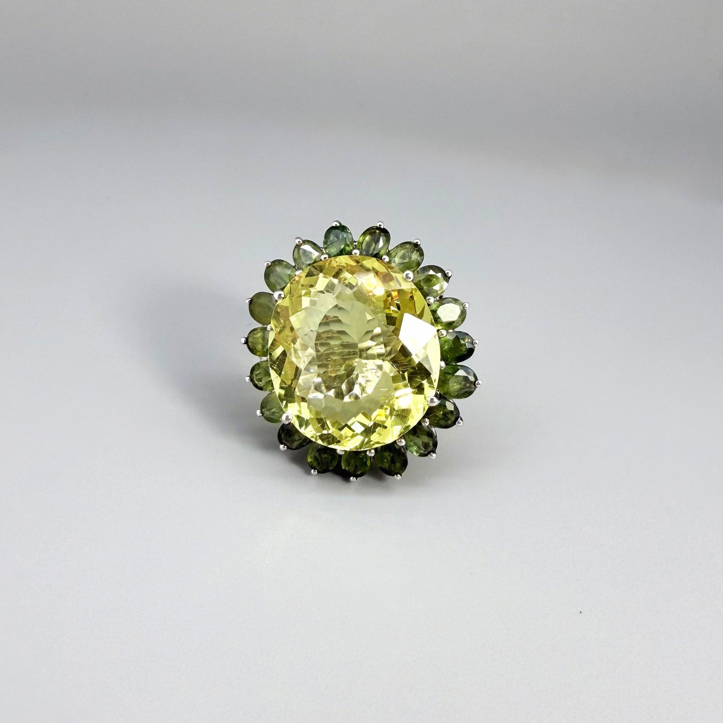 Statement lemon quartz ring in white gold plated sterling silver featuring a large yellow gemstone surrounded by green sapphires in a halo design.
