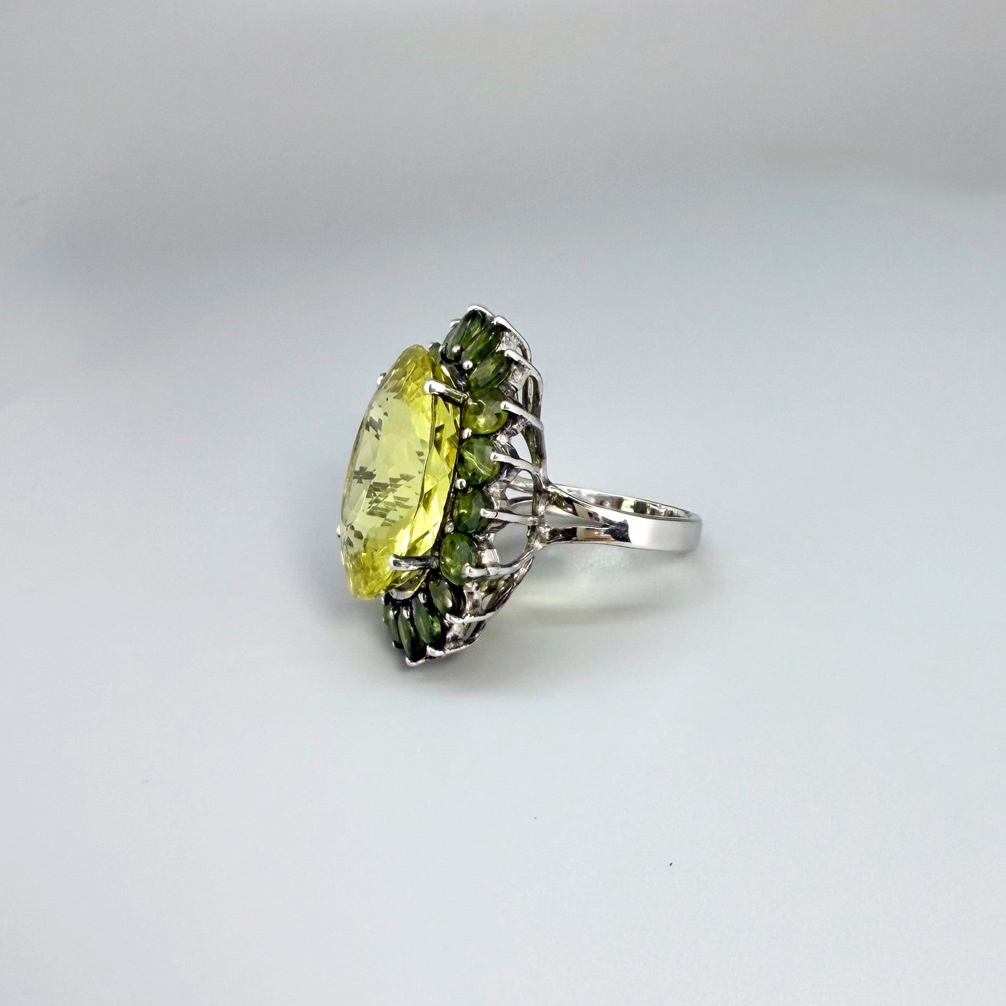 Statement lemon quartz ring in white gold plated sterling silver featuring a large yellow gemstone surrounded by green sapphires in a halo design.