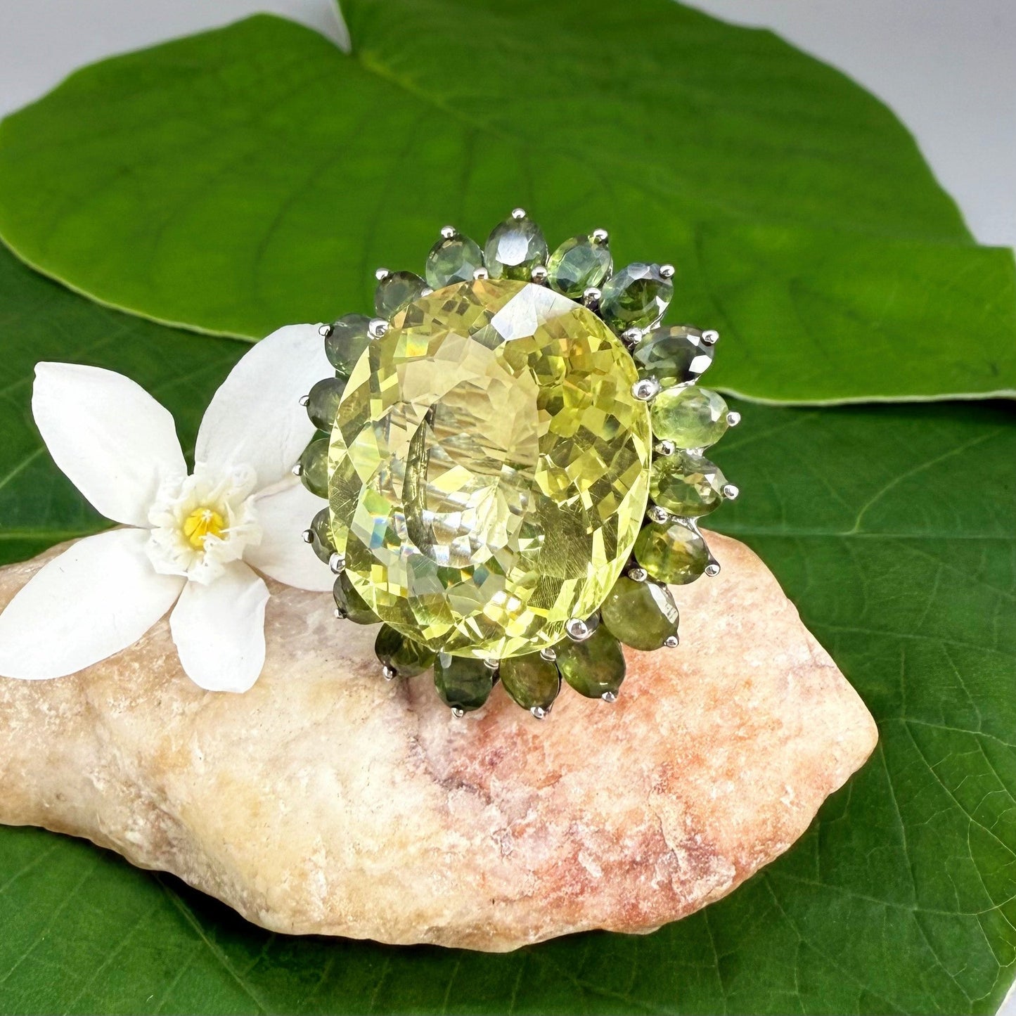 Statement lemon quartz ring in white gold plated sterling silver featuring a large yellow gemstone surrounded by green sapphires in a halo design.