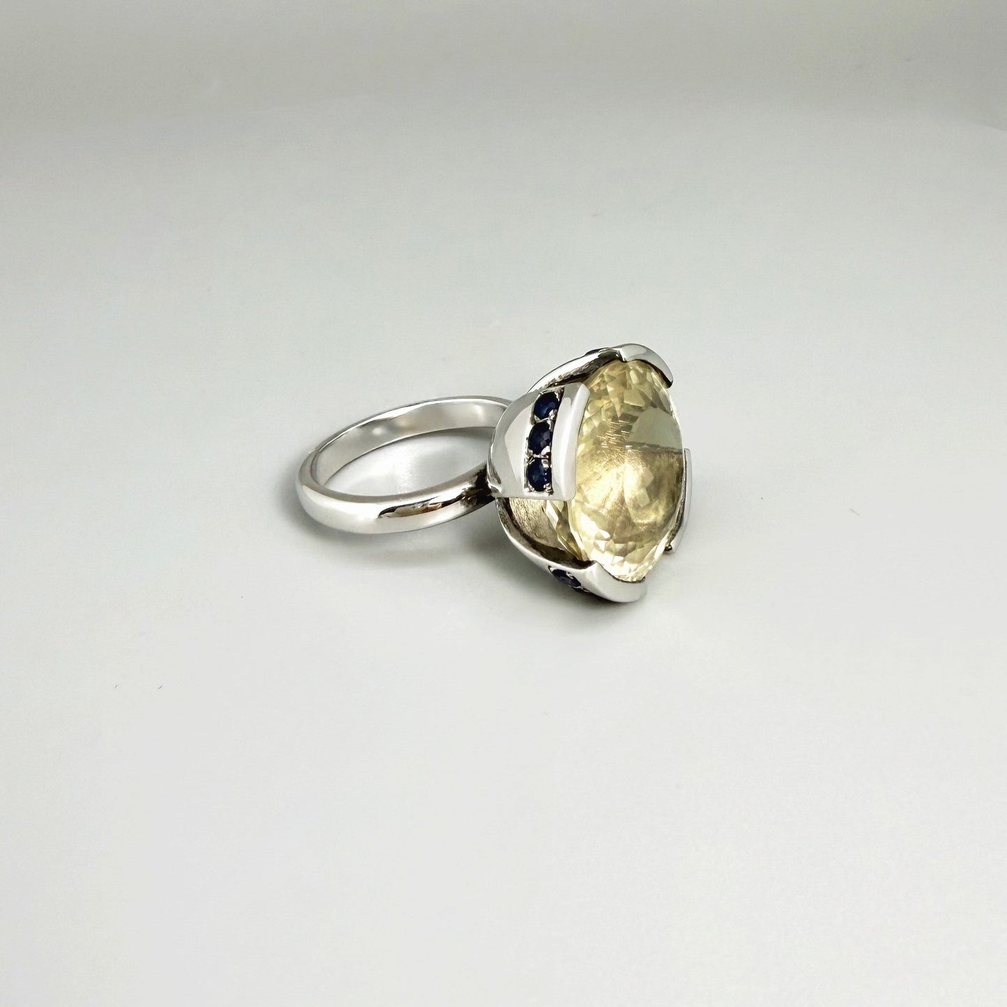 Lemon quartz and sapphire ring in white gold plated sterling silver, featuring a faceted yellow-green gemstone framed by deep blue sapphires, elegant and vibrant design.