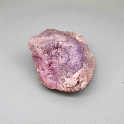 Natural ruby rough stone in lilac to lavender-pink tones, uncut corundum specimen photographed on neutral background.