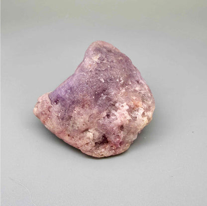Natural ruby rough stone in lilac to lavender-pink tones, uncut corundum specimen photographed on neutral background.