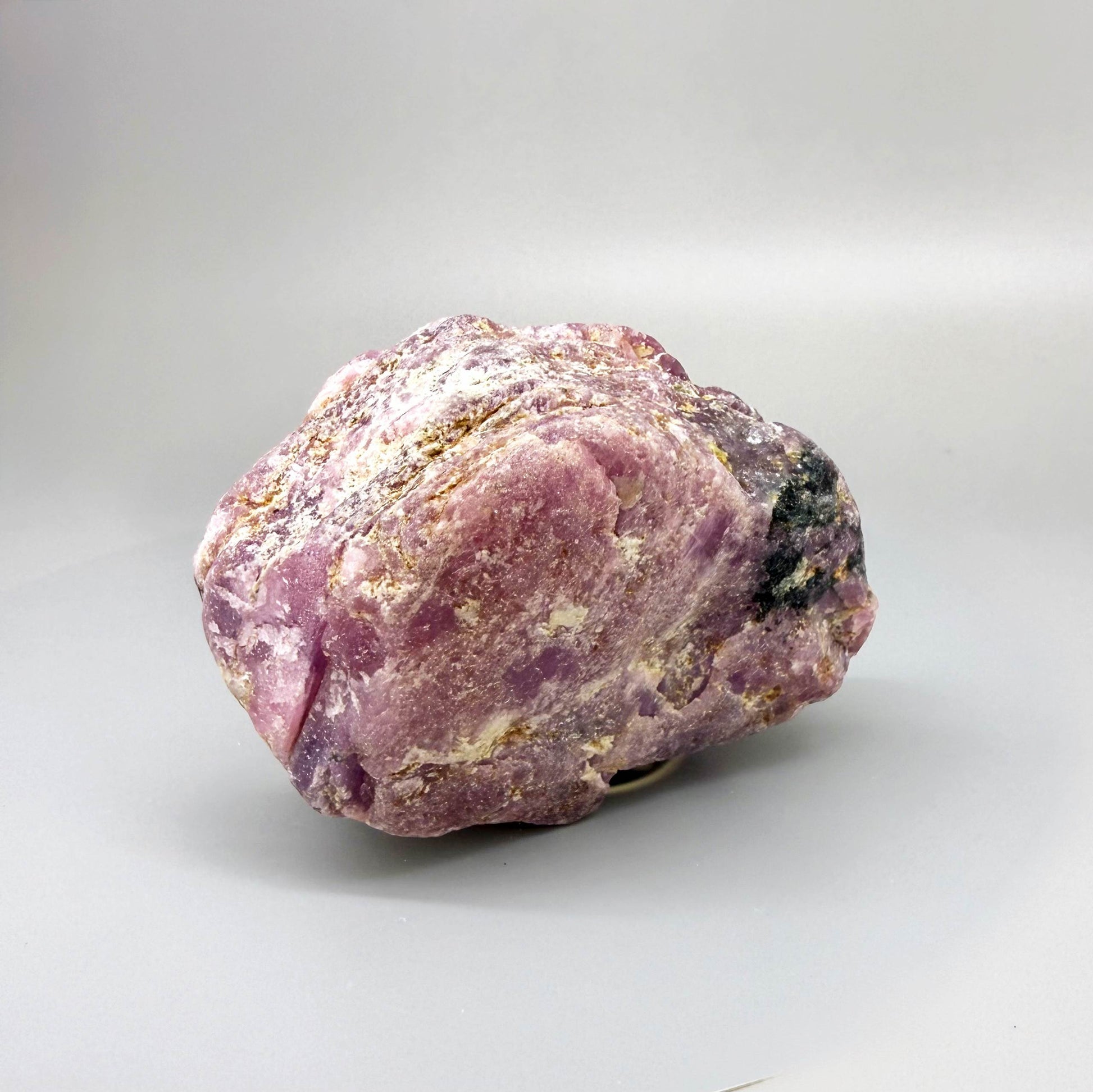 Natural rough ruby specimen with lilac to pinkish-violet tones in host rock, uncut and irregular, photographed on neutral background.