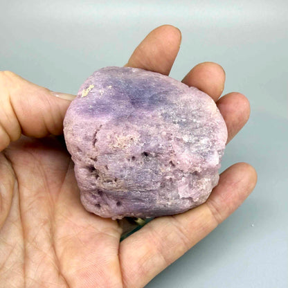Natural ruby rough stone in lilac to lavender-pink tones, uncut corundum specimen photographed on neutral background.