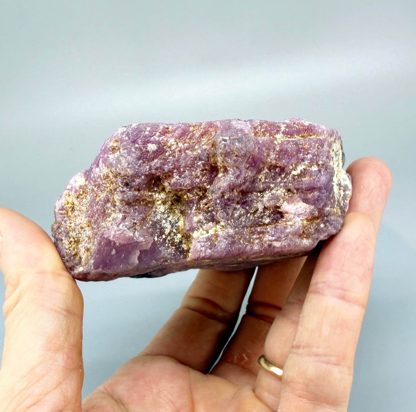 Natural rough ruby specimen with lilac to pinkish-violet tones in host rock, uncut and irregular, photographed on neutral background.