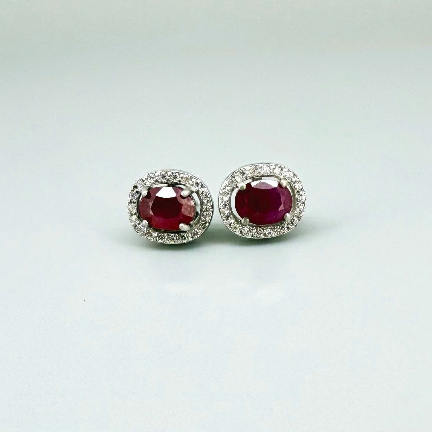 White gold plated sterling silver stud earrings with 2.6 ct natural rubies surrounded by cubic zirconia; classic halo design, elegant brilliance.