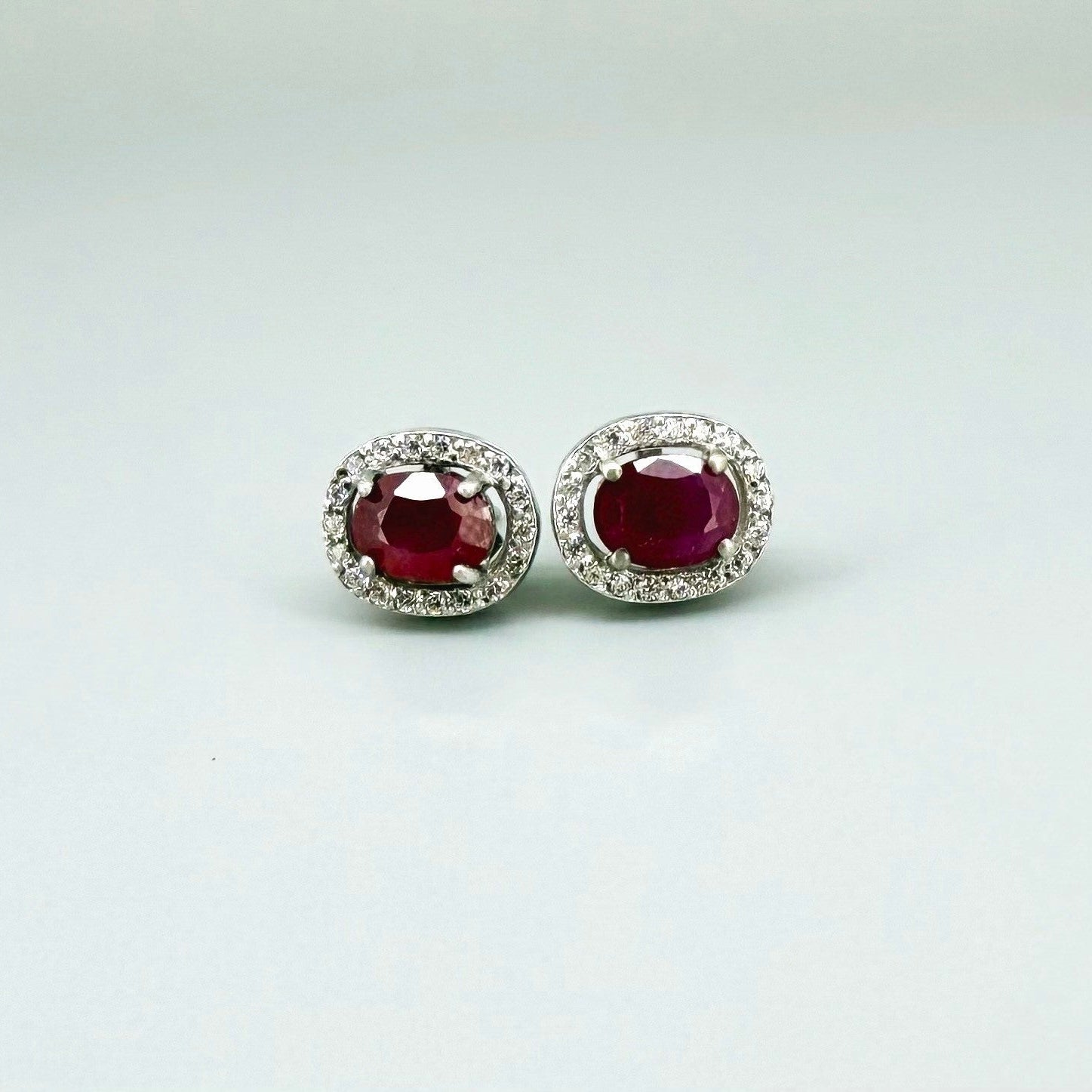 White gold plated sterling silver stud earrings with 2.6 ct natural rubies surrounded by cubic zirconia; classic halo design, elegant brilliance.