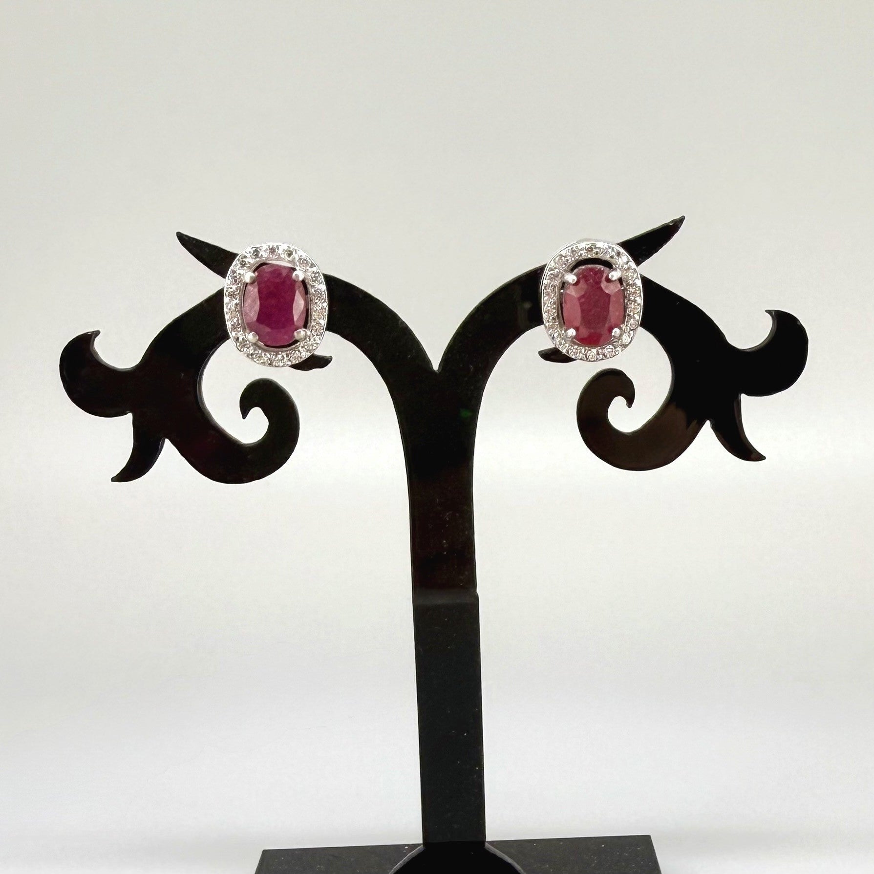 White gold plated sterling silver stud earrings with 2.6 ct natural rubies surrounded by cubic zirconia; classic halo design, elegant brilliance.