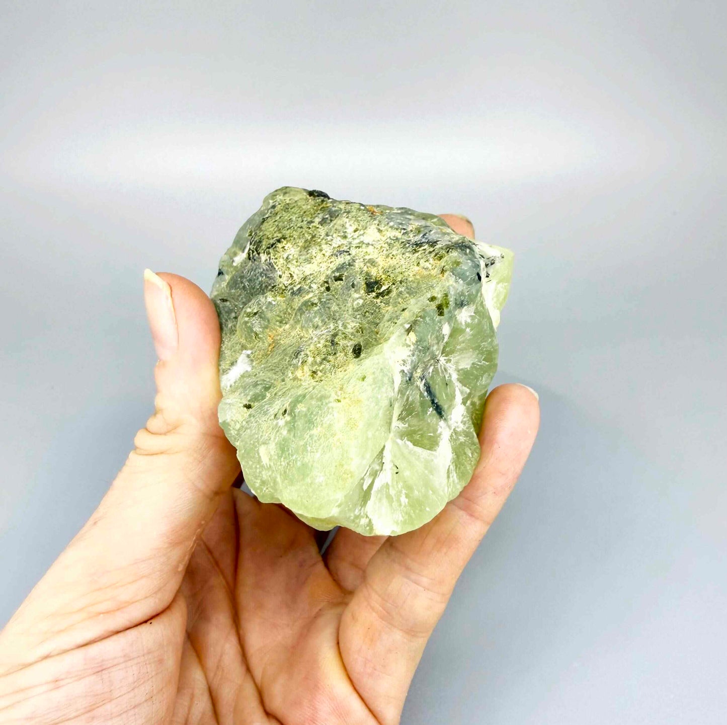 Prehnite rough stone in pale apple-green color with dark inclusions and semi-translucent body.
