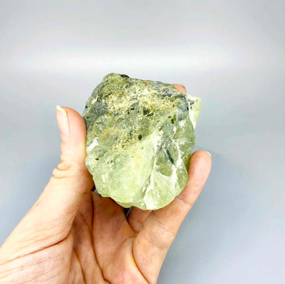 Prehnite rough stone in pale apple-green color with dark inclusions and semi-translucent body.