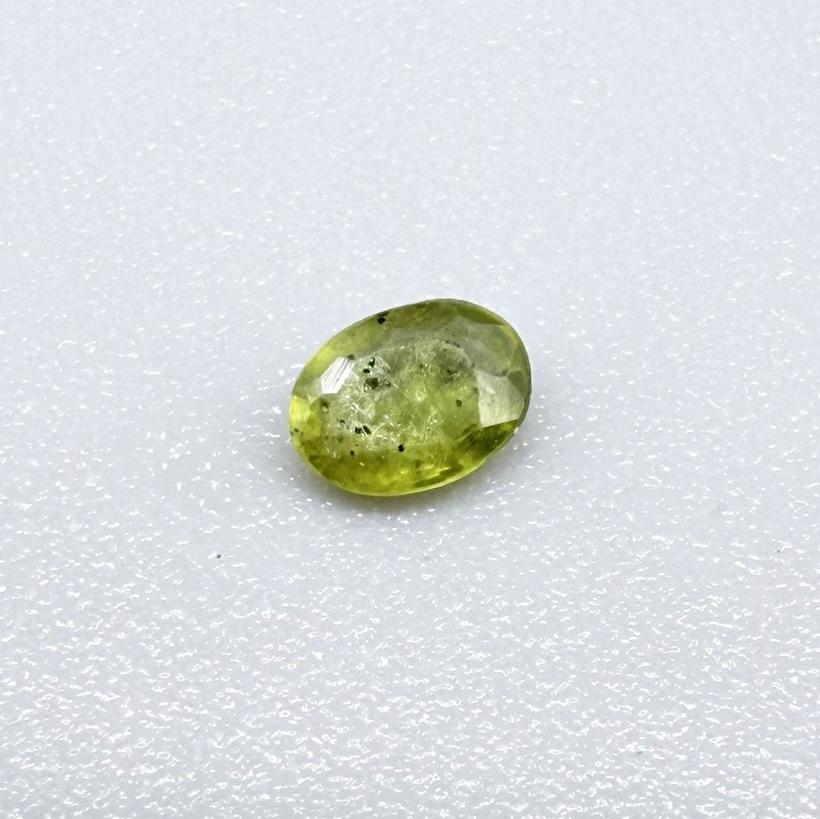 Oval yellow-green sapphire from Thailand, 0.21ct, unheated, with bright greenish-yellow color, visible dark natural inclusions and a transparent to slightly silky appearance.