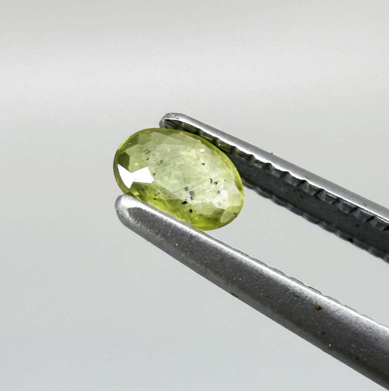Oval yellow-green sapphire from Thailand, 0.21ct, unheated, with bright greenish-yellow color, visible dark natural inclusions and a transparent to slightly silky appearance.