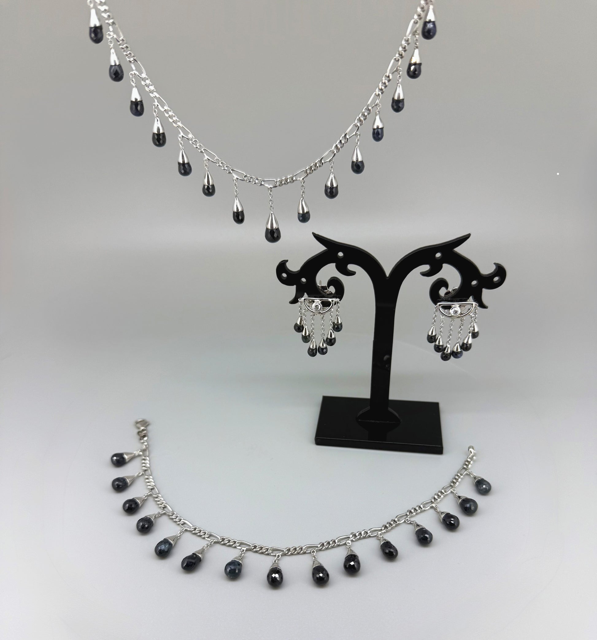 Necklace, bracelet and earrings set in white gold plated sterling silver with dark blue pineapple-cut sapphires, elegant jewelry displayed on neutral background.