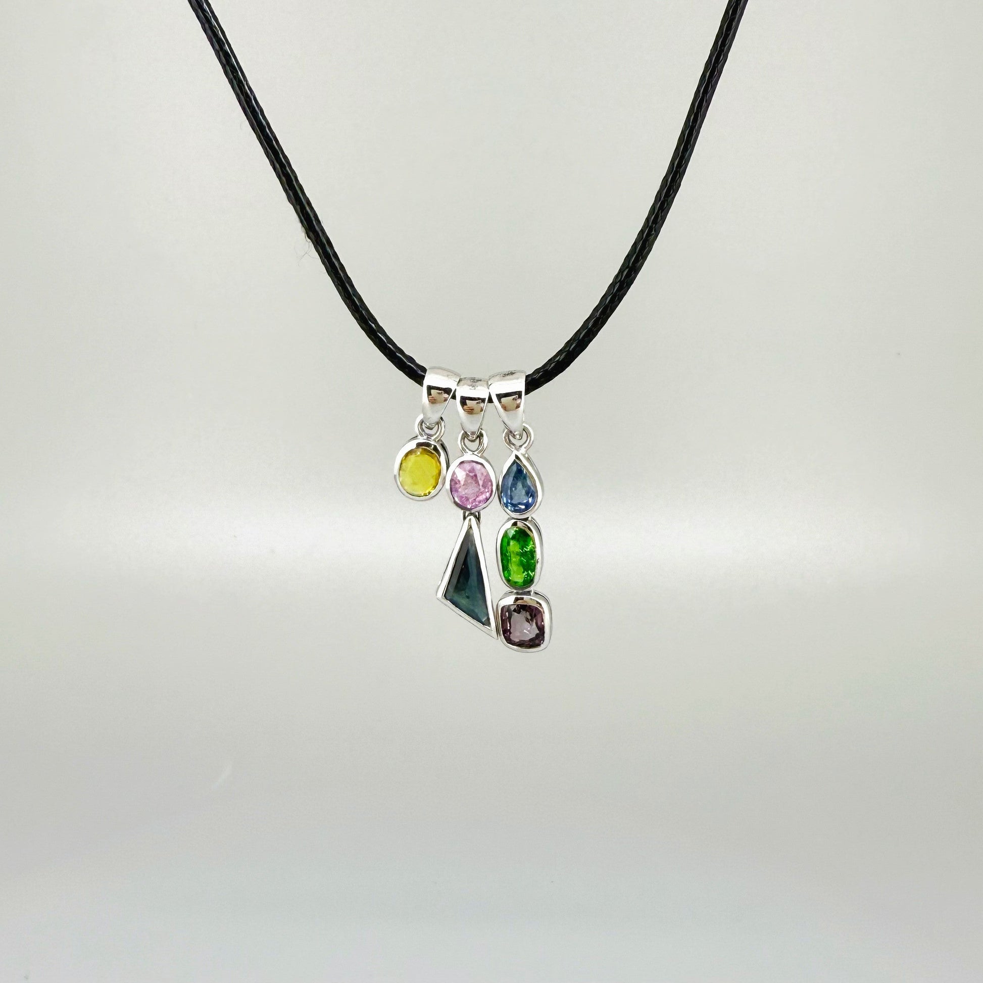 Handcrafted three-pendant gemstone necklace with sapphires, ruby, spinel, and tsavorite (5.81 ct total) on a black leather cord — modular design in white-gold-plated silver.
