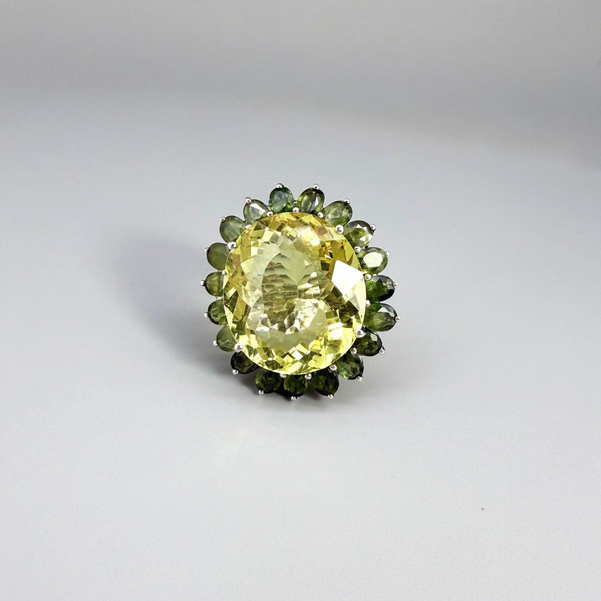 Statement lemon quartz ring in white gold plated sterling silver featuring a large yellow gemstone surrounded by green sapphires in a halo design.