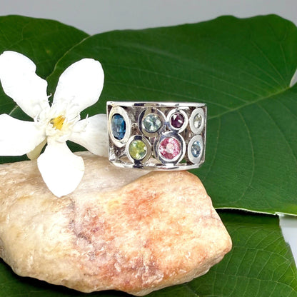 Multicolor sapphire band ring in white gold plated sterling silver, featuring faceted blue, yellow, pink, and green gemstones in elegant wide band design.